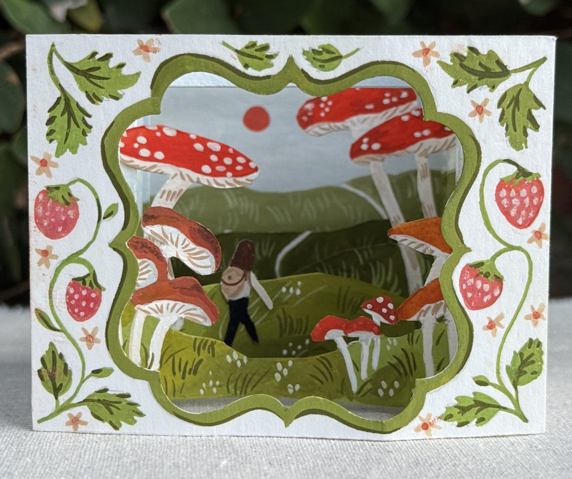 a small paper tunnel book depicting large mushrooms and a figure hiking through a landscape