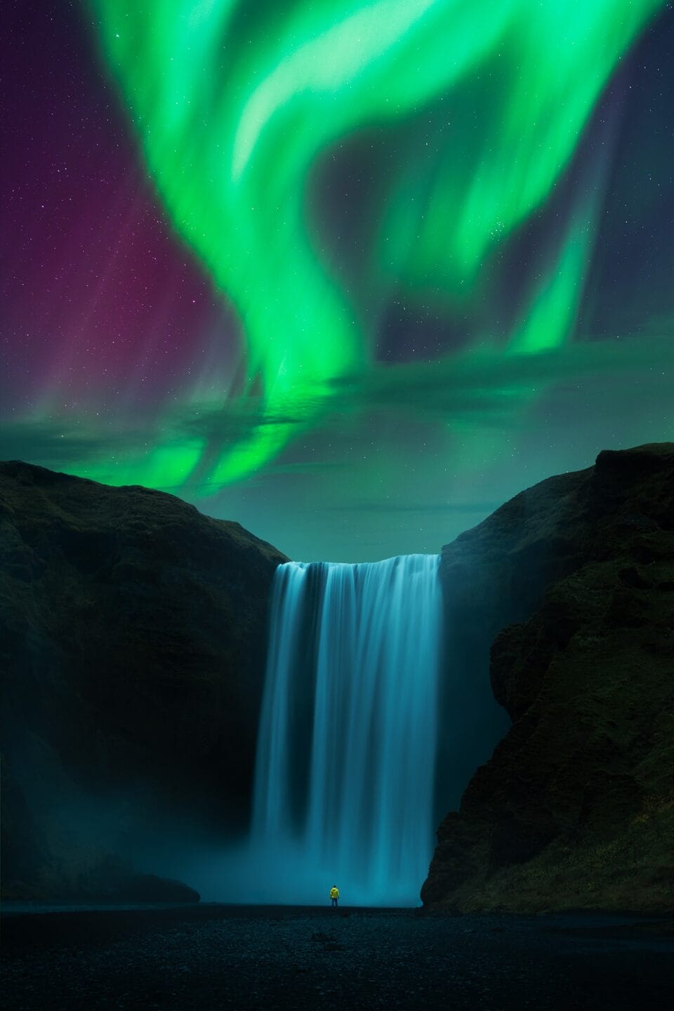 a small figure wearing a yellow jacket stands before a massive waterfall and a glowing sky displaying a monumental view of the Northern lights