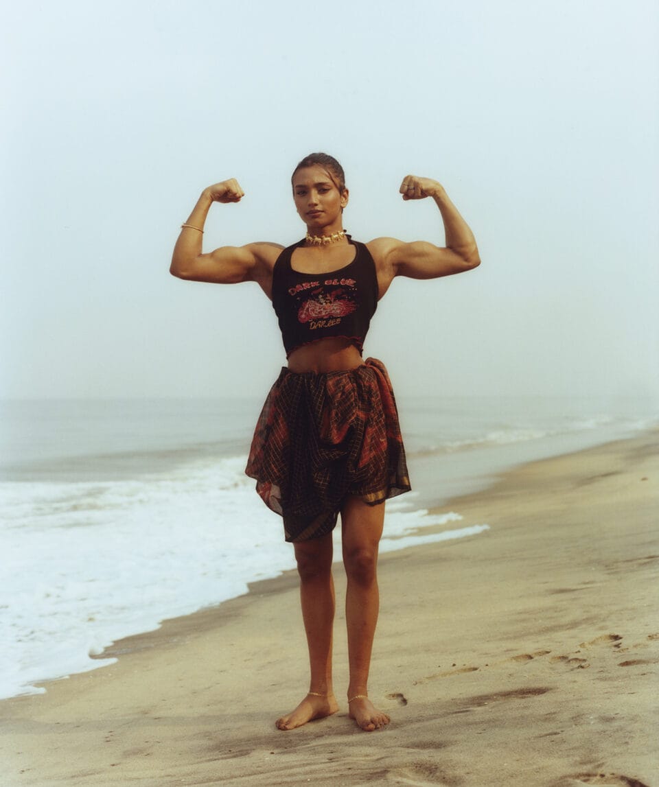 an indian woman in a skirt and tight black tank top flexes her arms on a beach