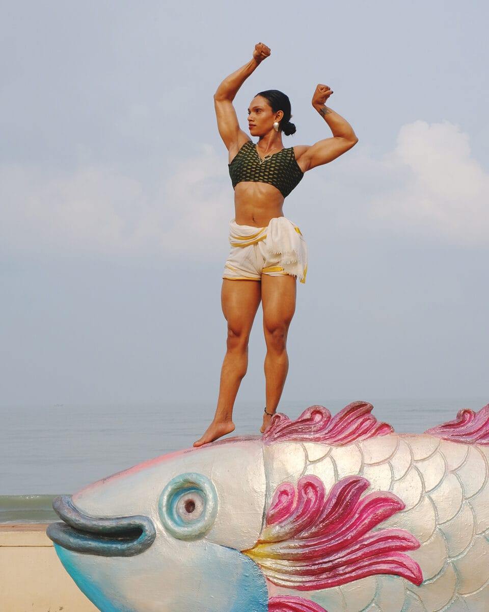 an indian woman in tight shorts and tank top stands atop a fish and flexes