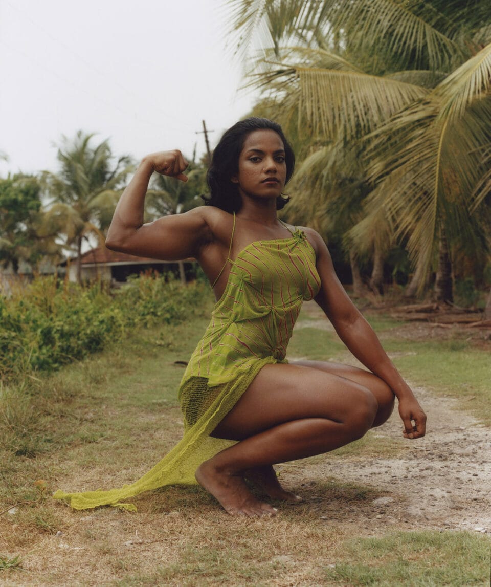 an indian woman in a green dress with large muscles squats down to flex her muscles in front of lush greenery